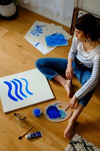 A woman is sitting on the floor painting stripes onto a canvas with blue paint.